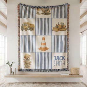Pu&ograve; includere: Una coperta tessuta con un motivo a griglia di strisce verticali blu e pannelli color crema. Il design include illustrazioni di un escavatore giallo, un camion con cassone ribaltabile, un bulldozer, un cono stradale e un orsacchiotto.