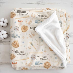May include: Cream-colored baby blanket with a baseball theme. The blanket features baseballs, bats, gloves, jerseys, and caps in shades of brown, blue, and white. The name "William Daniel" is printed on the blanket.