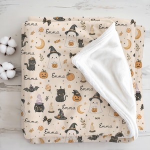 May include: A soft, folded blanket with a cream-colored background featuring a repeating Halloween pattern of ghosts in witch hats, black cats, pumpkins, bats, candles, moons, and bows, interspersed with the name Emma in black cursive script.