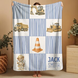 Pu&ograve; includere: Una coperta rettangolare con un motivo a griglia di strisce verticali blu e pannelli bianchi. I pannelli mostrano illustrazioni di un escavatore giallo, un camion con cassone ribaltabile, un bulldozer, un cono stradale arancione e il testo JACK SMITH.