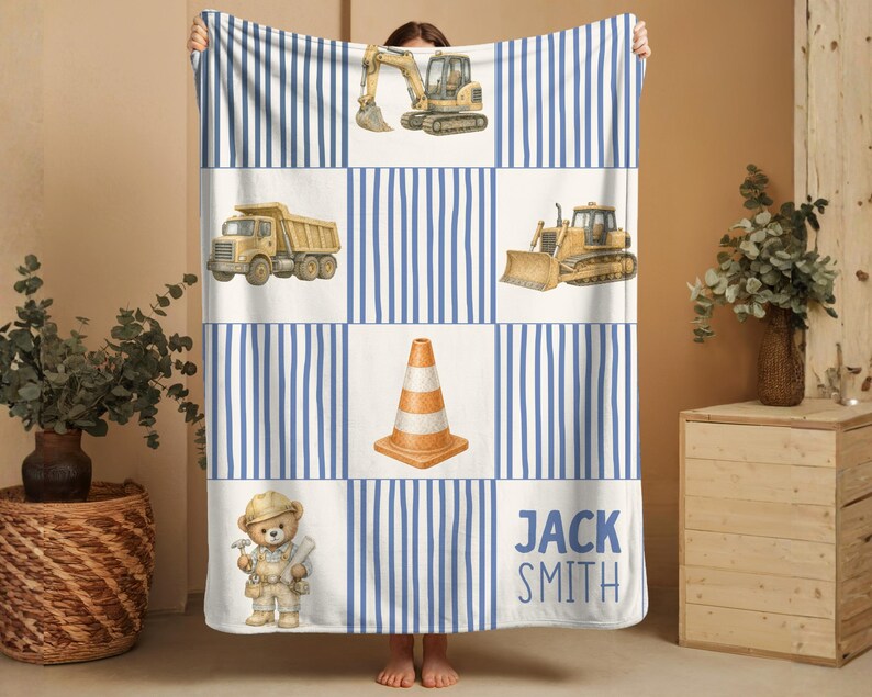 Pu&ograve; includere: Una coperta rettangolare con un motivo a griglia di strisce verticali blu e pannelli bianchi. I pannelli mostrano illustrazioni di un escavatore giallo, un camion con cassone ribaltabile, un bulldozer, un cono stradale arancione e il testo JACK SMITH.