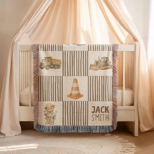 Pu&ograve; includere: Una coperta tessuta con un motivo a griglia con blocchi a righe alternati e illustrazioni di un camion con cassone ribaltabile giallo, un bulldozer giallo, un cono stradale arancione e un orsacchiotto con casco da cantiere, con il nome JACK SMITH.