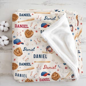 May include: A cream-colored baseball-themed blanket with the name "Daniel" in red and navy blue. The blanket features baseballs, bats, gloves, caps, and stars. The blanket is folded, revealing a soft, white interior.