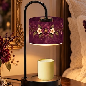 May include: A black metal candle warmer lamp featuring a cylindrical shade with a deep purple background, decorated with a floral pattern of purple and white flowers, positioned above a plain, light yellow cylindrical candle.