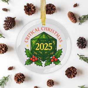 Puede incluir: Adorno navideño redondo y blanco con la inscripción "CRITICAL CHRISTMAS" en rojo. El adorno presenta un dado geométrico verde con el año "2025" en amarillo, rodeado de acebo y bayas. Se adjunta una cinta dorada para colgarlo.