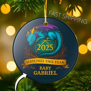 May include: A round, dark blue ceramic ornament with a teal dragon illustration. The dragon has purple wings and orange accents. Text includes "2025", "Hatched This Year", and "Baby Gabriel". A gold string is attached for hanging.