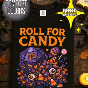 May include: Black t-shirt with the text "ROLL FOR CANDY" in orange. The graphic features a purple D20 die, candy, and eyeballs. The shirt has a round neck and is made of a soft material. The shirt is a Comfort Colors brand.