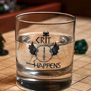 May include: A clear glass tumbler filled with liquid, featuring a black graphic design. The design includes the word "CRIT" above a circular emblem with wings and a sword, and the word "HAPPENS" below. The glass is on a patterned surface.