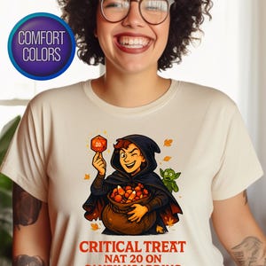 May include: Cream-colored t-shirt with a graphic of a character in a black cloak holding a dice and a bag of candy. Text reads "CRITICAL TREAT NAT 20 ON CANDY HOARDING". A green creature is also present.