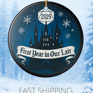 May include: A round, black Christmas ornament with a blue background featuring a castle silhouette, snowflakes, and the text "First Year in Our Lair" and "2025". A gold string is attached for hanging.