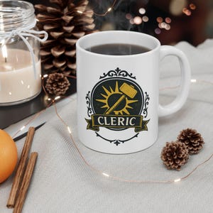 May include: White ceramic mug with a black and gold design featuring a sun, hammer, and the word "CLERIC" on a banner. The mug is filled with a dark liquid and has a curved handle.
