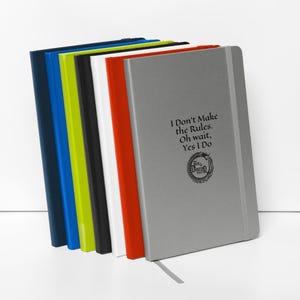 May include: A collection of hardcover notebooks in various colors, including navy blue, blue, lime green, black, white, red, and gray. The gray notebook features the text "I Don't Make the Rules. Oh wait, Yes I Do" and a circular logo.