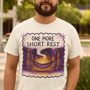 May include: White t-shirt with a graphic design featuring the text "ONE MORE SHORT REST". The design includes a hammock, pillars, and a starry night scene with a campfire. The shirt is being worn by a person.