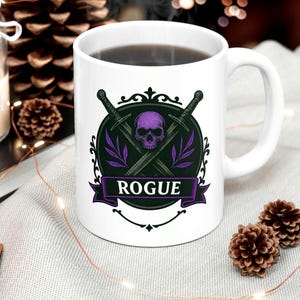 Può includere: Tazza in ceramica bianca con liquido nero all'interno. La tazza presenta un disegno con un teschio viola, spade incrociate e la parola "ROGUE" su uno stendardo. Il disegno è su uno sfondo nero e verde.