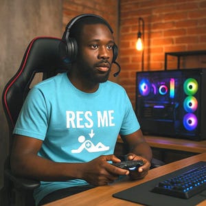 May include: A light blue t-shirt with the text "RES ME" and a graphic of a person lying down with a plus sign. The person is wearing a headset and holding a game controller. A computer with colourful lights is in the background.