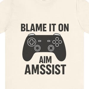 May include: Cream-colored t-shirt featuring a black graphic of a video game controller. The text "BLAME IT ON" is above the controller, and "AIM AMSSIST" is below. The shirt has a casual, relaxed fit.