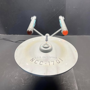 May include: A model of the U.S.S. Enterprise starship featuring a large circular saucer section with the text U.S.S. ENTERPRISE NCC-1701 printed on the top. The model includes two gray engine nacelles with glowing red and orange end caps.