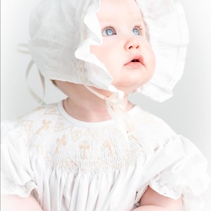 May include: A baby wearing a white bonnet with a ruffled edge and a matching dress. The dress features embroidered crosses and puffed sleeves. The baby has bright blue eyes and is looking upwards.