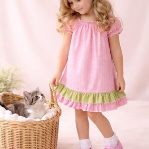 Pode incluir: Um vestido rosa com bolinhas e acabamento verde. O vestido é usado com sapatos rosa e meias brancas. Um gatinho está sentado em uma cesta de vime. A imagem inclui o texto "Juvie Moon designs".