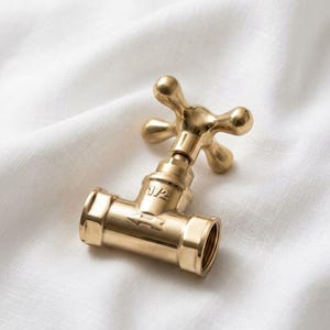 May include: A brass-colored faucet valve with a cross-shaped handle. The valve has a cylindrical body with a threaded end and an arrow indicating flow direction. The valve is set against a white, textured background.