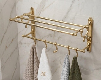May include: A gold-coloured towel rack with two horizontal bars and hooks for hanging towels. Four different coloured towels are hanging from the hooks. The towels are beige, white with an embroidered "M", grey, and olive green. The rack is mounted on a marble-patterned wall.