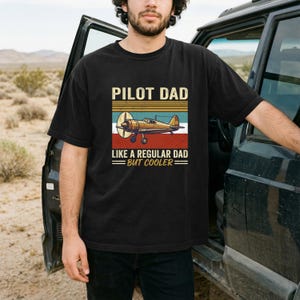 Könnte beinhalten: Schwarzes T-Shirt mit dem Text "PILOT DAD" über einer Grafik eines Oldtimer-Flugzeugs. Die Grafik hat ein rot-weiß-blaues Farbschema. Der Text "LIKE A REGULAR DAD BUT COOLER" steht unter der Flugzeuggrafik.