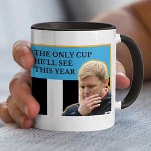 Puede incluir: Taza de cer&aacute;mica blanca con interior y asa negros. La taza presenta un rect&aacute;ngulo azul con el texto "THE ONLY CUP HE'LL SEE THIS YEAR" y una imagen de un hombre. El dise&ntilde;o incluye rayas blancas y negras.