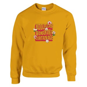 May include: Mustard yellow sweatshirt featuring the text "MIDDLE CLASS WOMAN OF A CERTAIN AGE" in a retro style. The text is in red, orange, and white, with daisy accents. The sweatshirt is a Gildan brand.