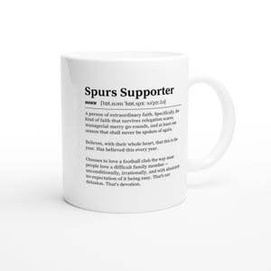 May include: White ceramic mug featuring the text "Spurs Supporter" and a definition. The definition describes a person of extraordinary faith in a football club. The mug has a curved handle.