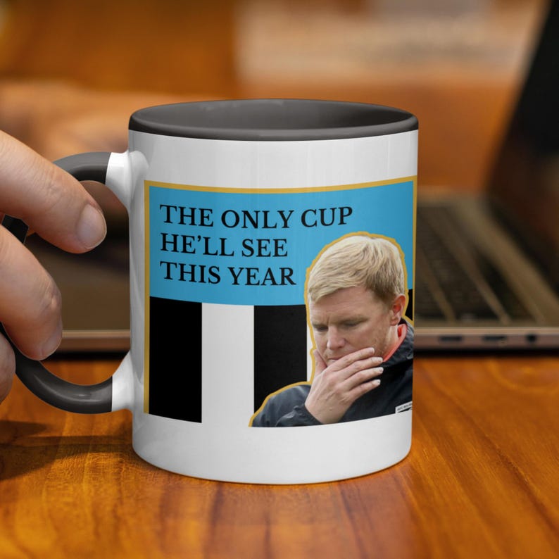 Puede incluir: Una taza de cer&aacute;mica blanca con interior y asa negros. La taza presenta un rect&aacute;ngulo azul con el texto "THE ONLY CUP HE'LL SEE THIS YEAR" y una imagen de un hombre. La taza est&aacute; sostenida por una persona.