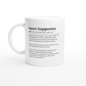 May include: White ceramic mug featuring the text "Spurs Supporter" and a definition of a fan. The definition describes a person of extraordinary faith. The mug has a curved handle and is white.