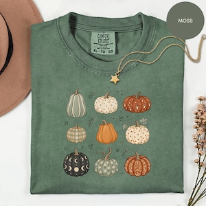 Könnte beinhalten: Ein moosgrünes T-Shirt mit einem Grafikdruck von neun Kürbissen in verschiedenen Farben und Mustern, darunter Karos, Punkte und florale Designs. Das T-Shirt hat einen Rundhalsausschnitt und ein Comfort Colors Markenetikett am Innenkragen.