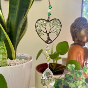 Puede incluir: Un atrapasoles en forma de corazón con un diseño de árbol, elaborado con alambre de cobre y cuentas verdes. Una lágrima de cristal transparente cuelga debajo, con las palabras "Made with Love". El atrapasoles está suspendido por una cadena de cobre.