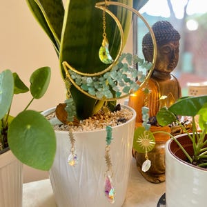 May include: A gold-coloured crescent moon and sun-shaped plant hanger with hanging crystals and green stones. The plant hanger is displayed in a white pot with a green plant. A small Buddha statue is in the background.