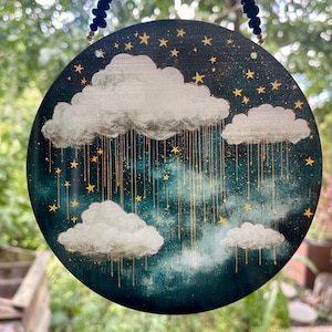 Puede incluir: Arte mural redondo de madera que representa una escena de cielo nocturno con nubes blancas, estrellas doradas y lluvia dorada. El fondo es de color verde azulado oscuro. El arte está suspendido por una cuerda con cuentas azules y blancas.
