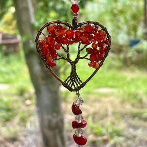 May include: A heart-shaped suncatcher with a wire tree design, filled with red stones. The tree is within a copper-coloured frame, with red and clear beads above and below. The background is a blurred view of greenery.