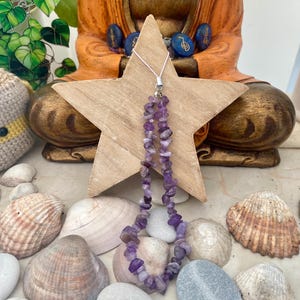 Puede incluir: Un collar hecho de pequeñas piedras irregulares de amatista púrpura. El collar cuelga frente a una estrella de madera. Conchas marinas y piedras lisas rodean el collar. Una estatua de Buda de madera está en el fondo.