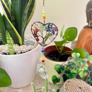 Puede incluir: Un atrapasoles en forma de corazón de color cobre con un diseño de árbol, lleno de piedras preciosas de colores que representan los chakras. Un prisma de cristal transparente cuelga debajo. El atrapasoles está suspendido por una cadena de cobre.