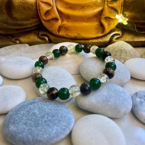 May include: A beaded bracelet featuring a mix of green, brown, and clear crystal beads. The bracelet is resting on smooth, gray and white stones. The background includes a golden Buddha statue and small fairy lights.