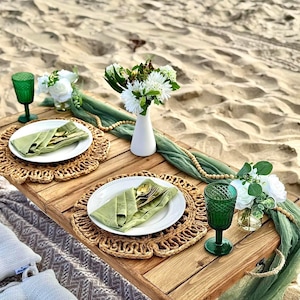 Peut inclure: Une table de pique-nique en bois pour deux personnes sur une plage de sable. La table est décorée de sets de table tressés, d'assiettes blanches, de serviettes vertes, de couverts dorés, de gobelets verts et de petits arrangements floraux blancs.