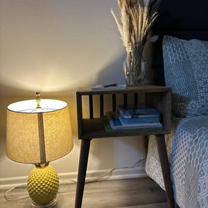 May include: A wooden end table with a shelf and four legs. The table has a vase with dried pampas grass on top and a stack of books on the shelf. The table is next to a bed with a gray and white patterned duvet cover.