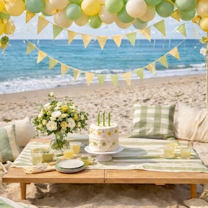 May include: A beach picnic scene with a decorated cake, flowers, and drinks. A wooden table is set with plates, glasses, and a cake on a stand. Balloons and bunting decorate the background, with the sea visible.