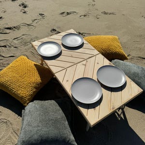 May include: A wooden picnic table with four gray plates and four cushions. Two of the cushions are yellow and two are gray. The table and cushions are on a sandy beach.