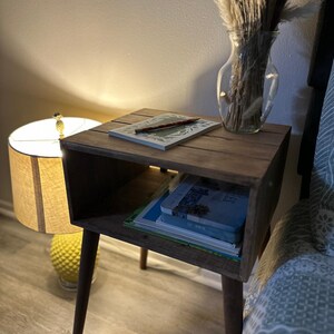 May include: A wooden nightstand with a shelf and a small drawer. The nightstand has a rustic finish and is painted brown. It has four legs and is sitting on a wooden floor. There is a lamp, a book, and a vase with dried flowers on top of the nightstand.