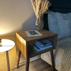 May include: A wooden end table with a shelf and tapered legs. The table has a natural wood finish and is decorated with a vase of dried pampas grass.