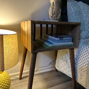 May include: A wooden end table with a shelf and four legs. The table has a rustic brown finish and is decorated with a vase of dried flowers. The table is next to a bed with a patterned duvet cover.
