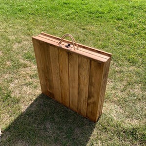 Puede incluir: Una mesa plegable de madera con un asa de cuerda. La mesa está cerrada y está sobre un césped.