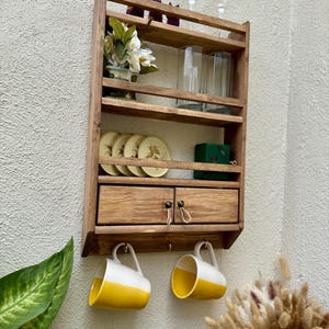 Peut inclure: Étagère murale en bois avec plusieurs niveaux. Elle comprend deux petits tiroirs, des crochets pour tasses et expose des assiettes, des verres et des objets décoratifs. Deux tasses jaunes et blanches sont suspendues en dessous.