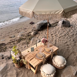 May include: A beach picnic setup featuring a beige fringed umbrella, a wooden picnic table, and two round floor cushions. The table is set with plates, fans, and candles, with a floral arrangement in a woven basket. The scene is on a sandy beach.