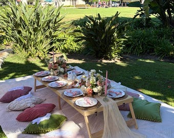 May include: A picnic setup on a sunny day, featuring a low wooden table set with plates, flowers, and candles. Pillows in various colors surround the table on a white quilted blanket. A fringed parasol provides shade.